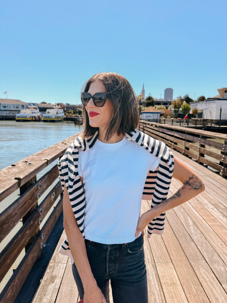 Person wearing a red purse, white t-shirt, and striped sweater standing on a pier in San Francisco, with the waterfront in the background.