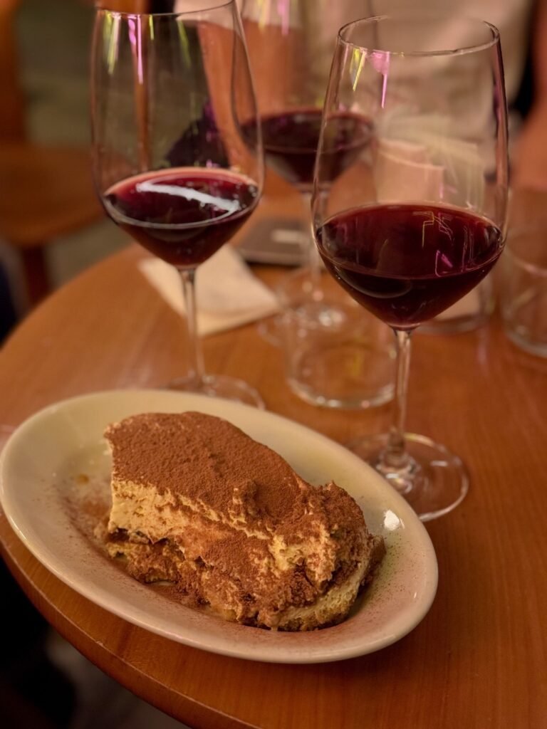 A close-up of a rich and creamy tiramisu dusted with cocoa powder, plus a glass of wine served in a cozy Zurich café