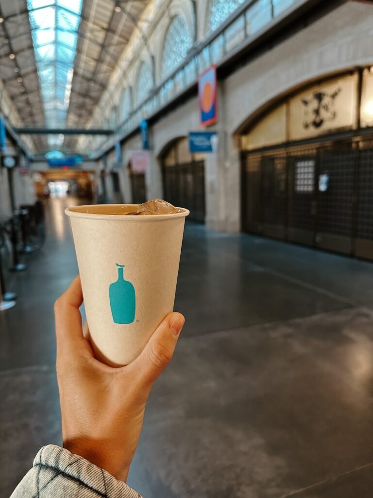 A hand holding a refreshing Blue Bottle iced coffee inside the Ferry Building, with the market’s bustling atmosphere and artisan vendors in the background.