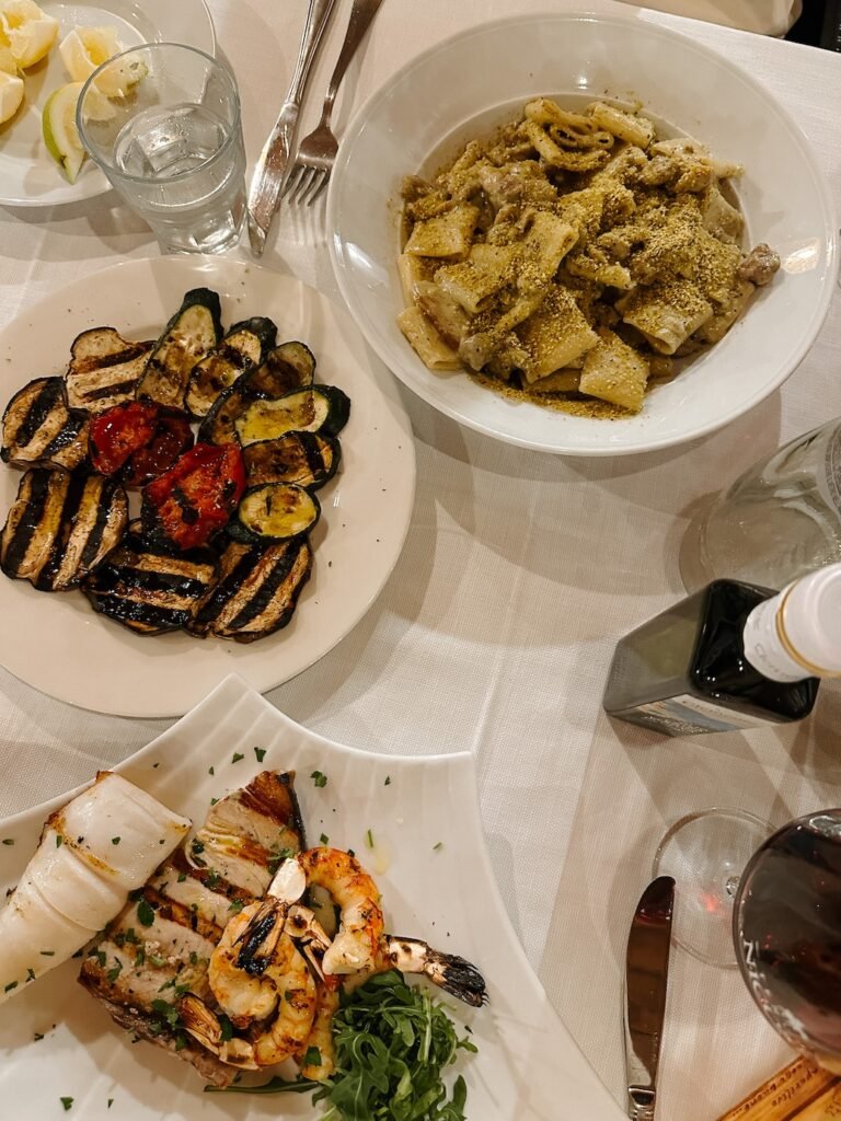 A table setting with grilled vegetables, seafood skewers with prawns and fish, and a bowl of rigatoni pasta topped with sausage and a green pistachio dust, served with a glass of red wine.