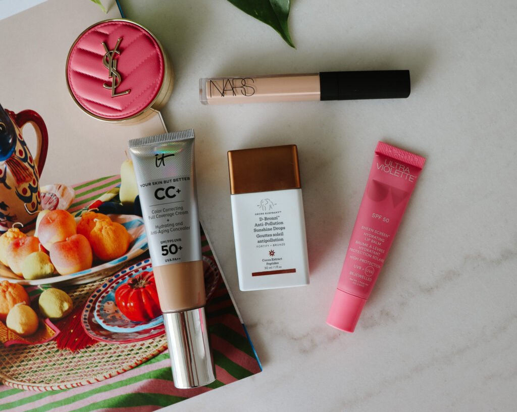 Flat lay of summer travel beauty essentials on a marble tabletop: CC+ cream SPF 50, pink SPF 50 lip balm, NARS concealer, and anti-pollution bronzing drops, surrounded by a decorative magazine of a bowl of fruit and greenery.
