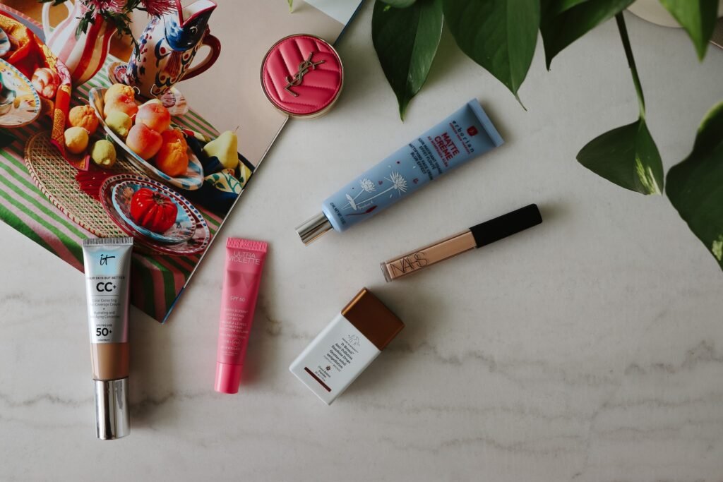 Flat lay of summer travel beauty essentials on a marble tabletop: CC+ cream SPF 50, pink SPF 50 lip balm, floral-scented mattifying face cream, NARS concealer, and anti-pollution sunscreen drops, surrounded by a decorative bowl of fruit and greenery.