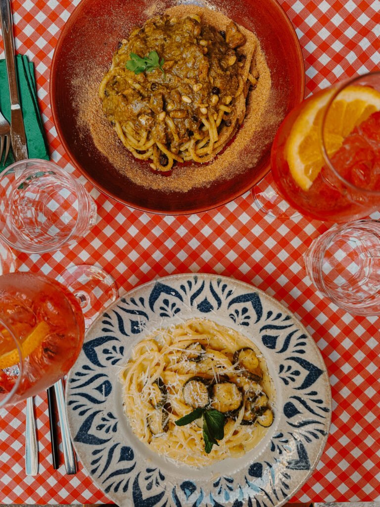 Two plates of Italian pasta dishes served on a red gingham tablecloth with glasses of Aperol Spritz and water; one pasta is topped with a rich meat sauce and pine nuts, and the other is a creamy pasta with zucchini and cheese.